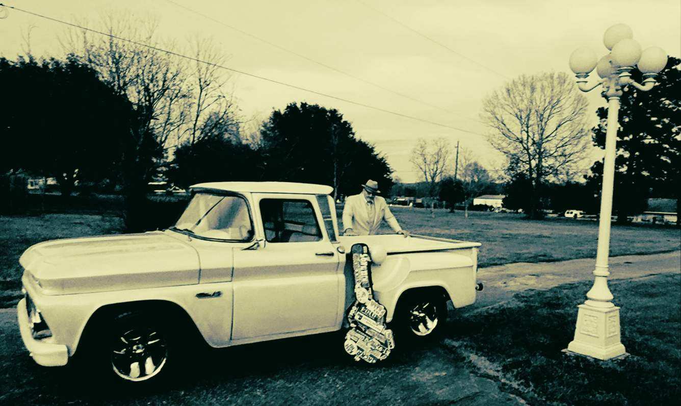Uncle Ryano standing behind a classic, white pickup truck with his guitar case in front.